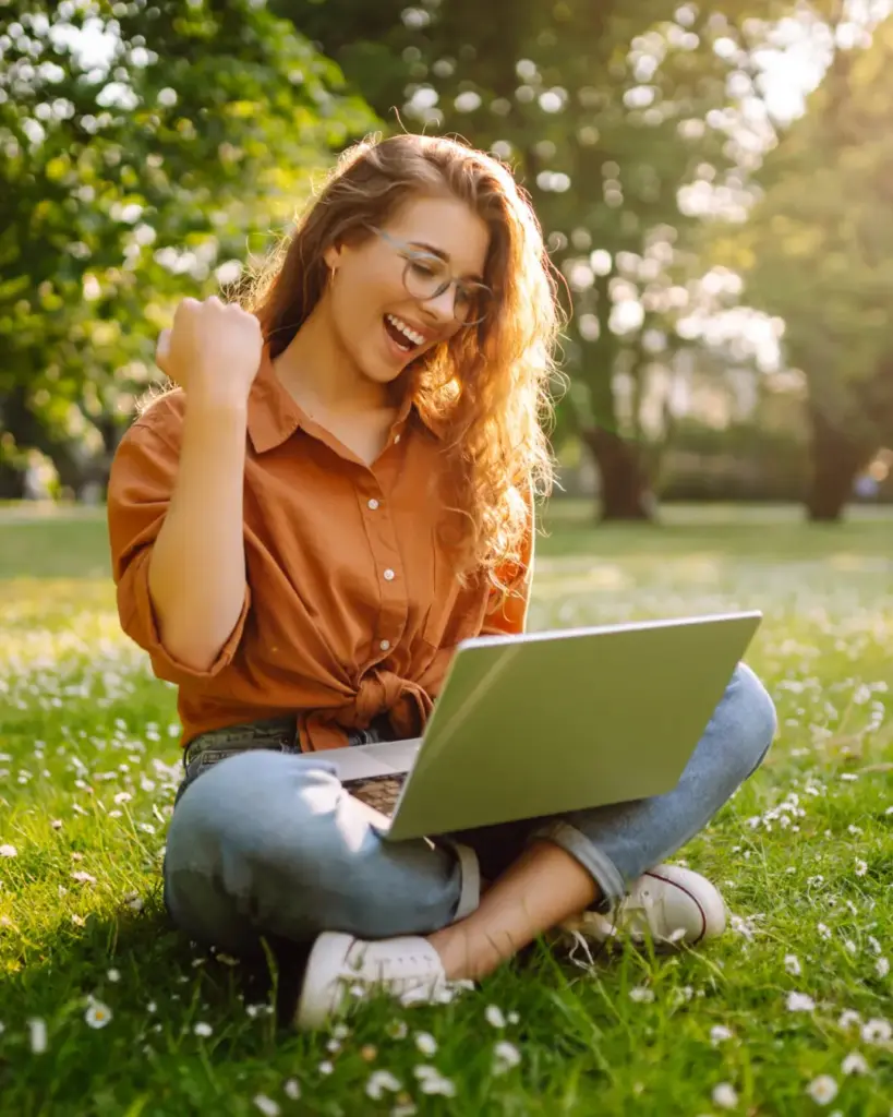 Sinnbild: 1 zu 1 Coaching um Karrierweg als Frau zu finden und endlich glücklich zu sein. Bild einer jungen, blnden Frau mit langen welligen Haaren. Sie sitzt im Schneidersitz bei Sonnenschein in einem Park auf einer Wiese. Sie jubelt und hat einen Laptop auf dem Schoss.